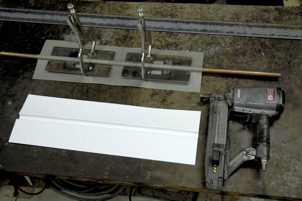 This picture shows a piece of grooved flashing (white, upside down), a piece of right side up grooved flashing (gray) mounted over a copper pipe with the edge clamps in place (note holes in them to