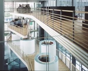 by a striking foyer building which houses the entrance area The symmetry is broken, however, by the presence of three floating, curvilinear overhanging balconies in the transparent foyer volume,