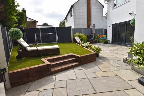 To one side of the driveway is a raised chipped slate area adjoined by a further raised artificial lawned area.